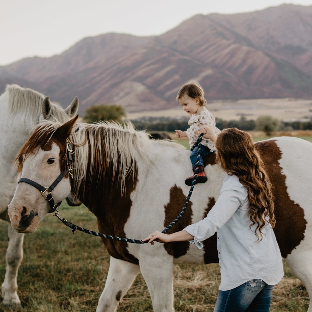toddler on a horse as her mother helps lead the horse through a field