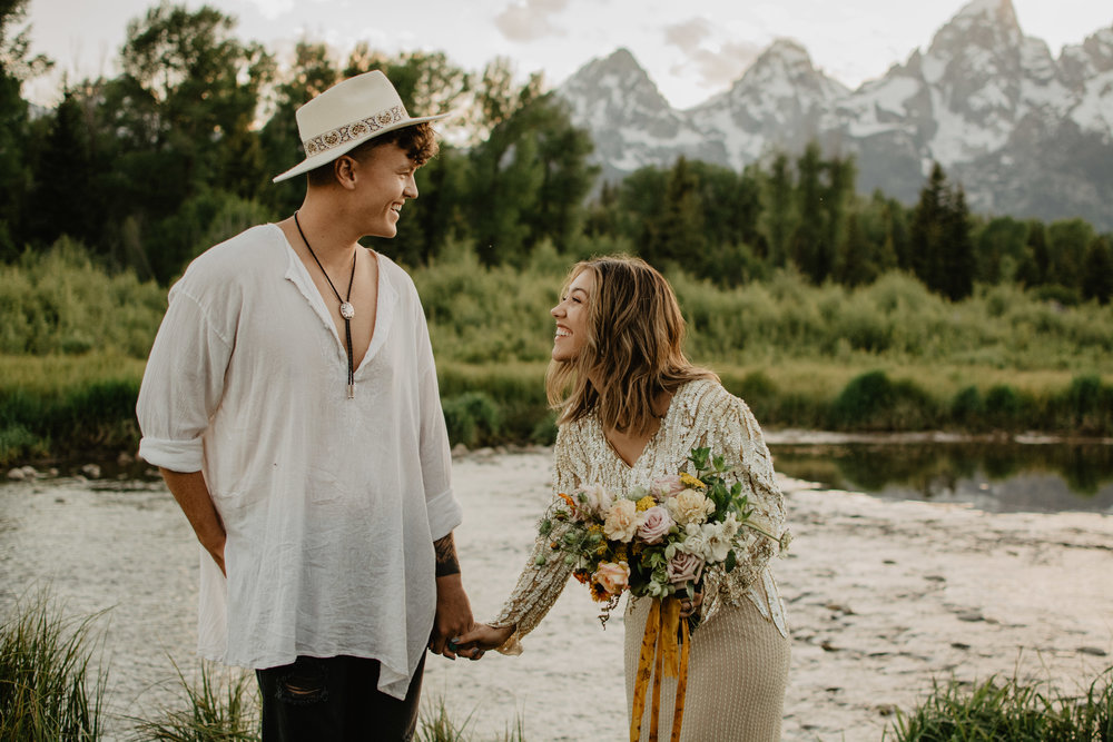 bride and groom in a elopement location in Jackson Hole and the Grand Tetons standing infront of a river with the Tetons in the background as they laugh and smile with one another on their wedding day