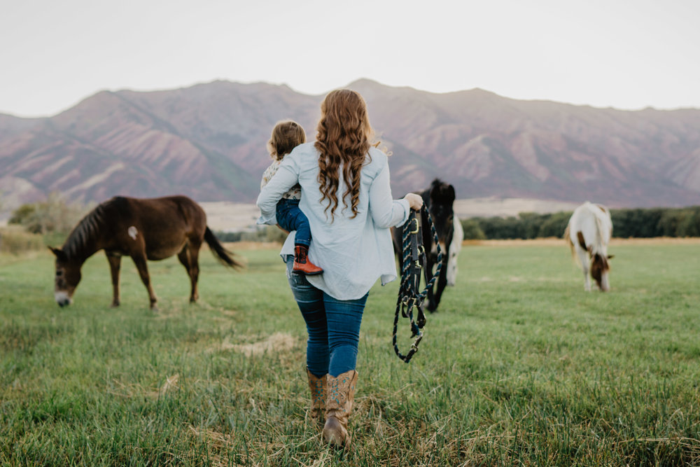 mother holding small child on her hip as she walks into a pasture with horses on how to run your business and be a mom