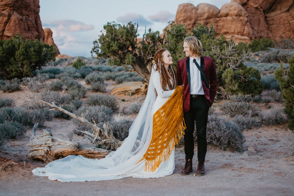 boho bride with dreads and an orange shaw holds onto her groom who is in a red suit coat as they stand in Moab Utah's red rocks for their adventure elopement wedding day Jocilyn Bennett Photography | How to Plan an Elopement | Why Elope | Reasons to Elope | Destination Wedding Photographer | Utah Wedding Photographer | Adventure Photographer | Destination Elopement Photographer | Traveling Photographer | Arches National Park | Arches National Park Elopement | Southern Utah Elopement | Boho Bride | Bride with dreads |