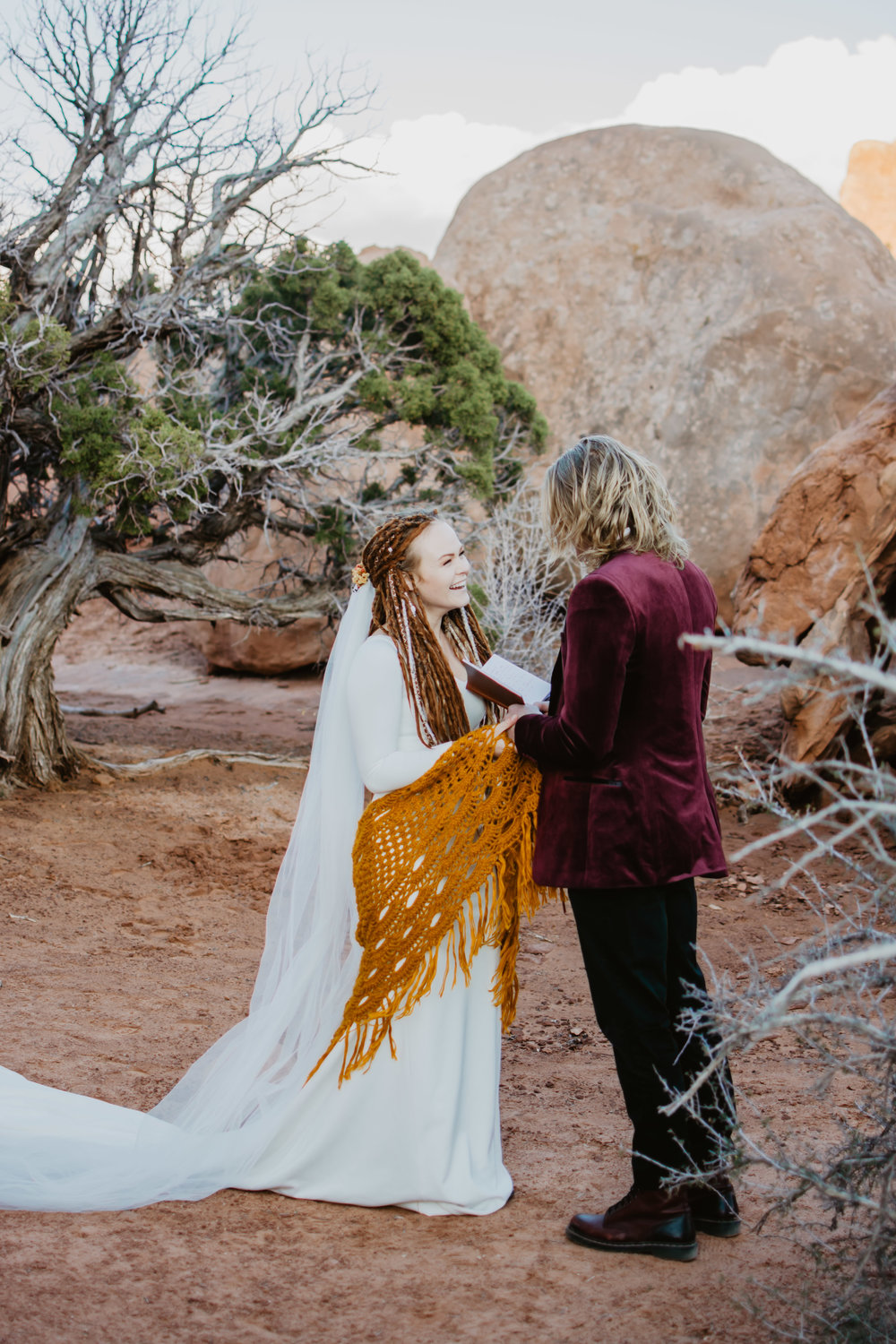 beautiful bride in dread locks and a simple white wedding dress with a burnt orange shaw smiles as her groom read his vows to her as they stand in the desert in Utah on their wedding day Jocilyn Bennett Photography | How to Plan an Elopement | Why Elope | Reasons to Elope | Destination Wedding Photographer | Utah Wedding Photographer | Adventure Photographer | Destination Elopement Photographer | Traveling Photographer | Arches National Park | Arches National Park Elopement | Southern Utah Elopement | Boho Bride | Bride with dreads |