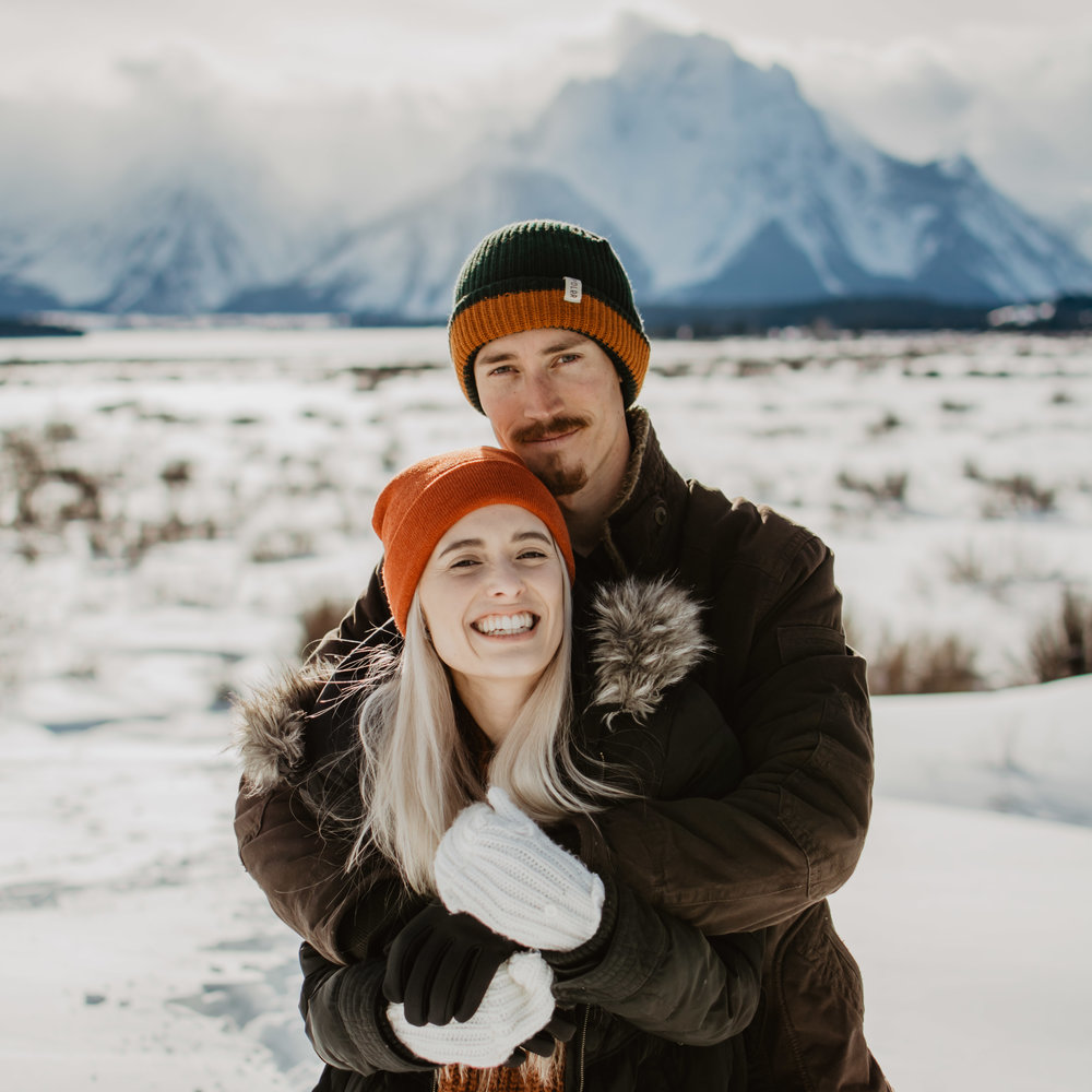 Man standing behind a woman and holding her as they smile at the camera for their engagement photos in the Grand Tetons in the winter | Jocilyn Bennett Photography | Winter Engagements | Snowy Engagements | Wyoming Engagement Photography | Utah Wedding Photographer | Winter Engagement Poses | What to Wear for Winter Engagements | Destination Wedding Photographer | Outfit ideas for Winter Engagements | Adventure Photographer | Wyoming Photographer | Winter Engagements in Jackson Hole | Jackson Hole Engagements | Wyoming Bride | Wyoming Wedding Photographer | Jackson Hole Wedding Photographer |