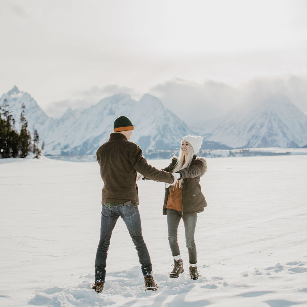 Man and woman dancing in the snow in front of the Tetons while wearing hats and coats for their winter engagement session | Jocilyn Bennett Photography | Winter Engagements | Snowy Engagements | Wyoming Engagement Photography | Utah Wedding Photographer | Winter Engagement Poses | What to Wear for Winter Engagements | Destination Wedding Photographer | Outfit ideas for Winter Engagements | Adventure Photographer | Wyoming Photographer | Winter Engagements in Jackson Hole | Jackson Hole Engagements | Wyoming Bride | Wyoming Wedding Photographer | Jackson Hole Wedding Photographer |