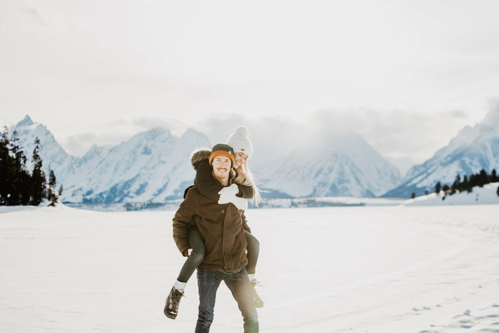 Winter engagemetn session in the Grand Tetons with man giving woman a piggyback ride through the snow in front of the Tetons | Jocilyn Bennett Photography | Winter Engagements | Snowy Engagements | Wyoming Engagement Photography | Utah Wedding Photographer | Winter Engagement Poses | What to Wear for Winter Engagements | Destination Wedding Photographer | Outfit ideas for Winter Engagements | Adventure Photographer | Wyoming Photographer | Winter Engagements in Jackson Hole | Jackson Hole Engagements | Wyoming Bride | Wyoming Wedding Photographer | Jackson Hole Wedding Photographer |