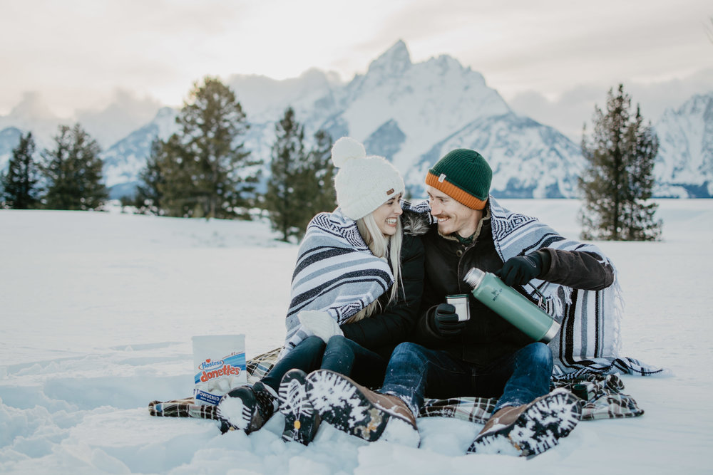 man and woman snuggles up in a wool tribal print blanket while sitting in the snow and holding thermoses in front of the Grand Tetons mountain range |Jocilyn Bennett Photography | Winter Engagements | Snowy Engagements | Wyoming Engagement Photography | Utah Wedding Photographer | Winter Engagement Poses | What to Wear for Winter Engagements | Destination Wedding Photographer | Outfit ideas for Winter Engagements | Adventure Photographer | Wyoming Photographer | Winter Engagements in Jackson Hole | Jackson Hole Engagements | Wyoming Bride | Wyoming Wedding Photographer | Jackson Hole Wedding Photographer |