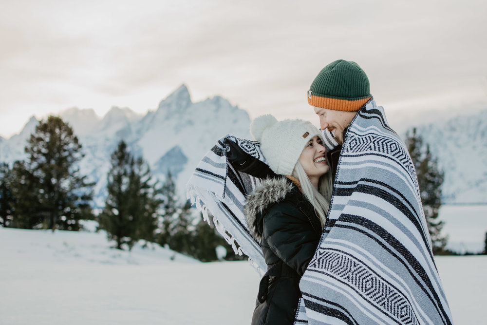 man wrapping up woman in a blanket as they stand in the snow with trees and the Grand Tetons in the background | Jocilyn Bennett Photography | Winter Engagements | Snowy Engagements | Wyoming Engagement Photography | Utah Wedding Photographer | Winter Engagement Poses | What to Wear for Winter Engagements | Destination Wedding Photographer | Outfit ideas for Winter Engagements | Adventure Photographer | Wyoming Photographer | Winter Engagements in Jackson Hole | Jackson Hole Engagements | Wyoming Bride | Wyoming Wedding Photographer | Jackson Hole Wedding Photographer |
