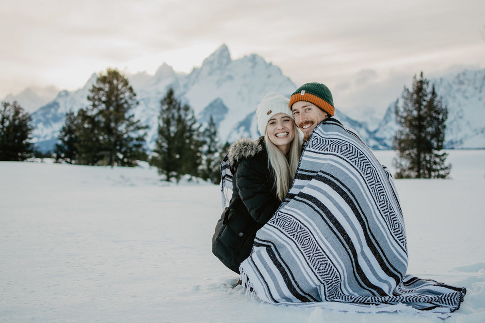 Grand Teton winter engagemnt session with man and woman crouched over with a blanket to share | Jocilyn Bennett Photography | Winter Engagements | Snowy Engagements | Wyoming Engagement Photography | Utah Wedding Photographer | Winter Engagement Poses | What to Wear for Winter Engagements | Destination Wedding Photographer | Outfit ideas for Winter Engagements | Adventure Photographer | Wyoming Photographer | Winter Engagements in Jackson Hole | Jackson Hole Engagements | Wyoming Bride | Wyoming Wedding Photographer | Jackson Hole Wedding Photographer |