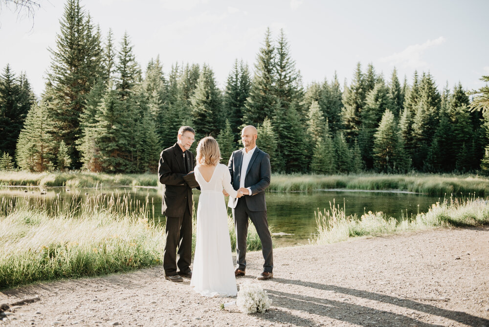Grand Teton National Park adventure elopement with mand and woman holding hands as to officiant marries the two in front of a river and tall pine trees Jocilyn Bennett Photography | How to Plan an Elopement | Why Elope | Reasons to Elope | Destination Wedding Photographer | Utah Wedding Photographer | Adventure Photographer | Destination Elopement Photographer | Traveling Photographer | Grand Teton Elopement | Grand Teton Photographer | National Park Weddings | Mountain elopement |