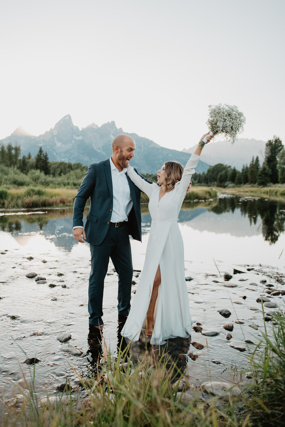 bride and groom standing in a rocky stream together in front of the Tetons as they smile at each other with the brides bouquet in the air sJocilyn Bennett Photography | How to Plan an Elopement | Why Elope | Reasons to Elope | Destination Wedding Photographer | Utah Wedding Photographer | Adventure Photographer | Destination Elopement Photographer | Traveling Photographer | Grand Teton Elopement | Grand Teton Photographer | National Park Weddings | Mountain elopement |