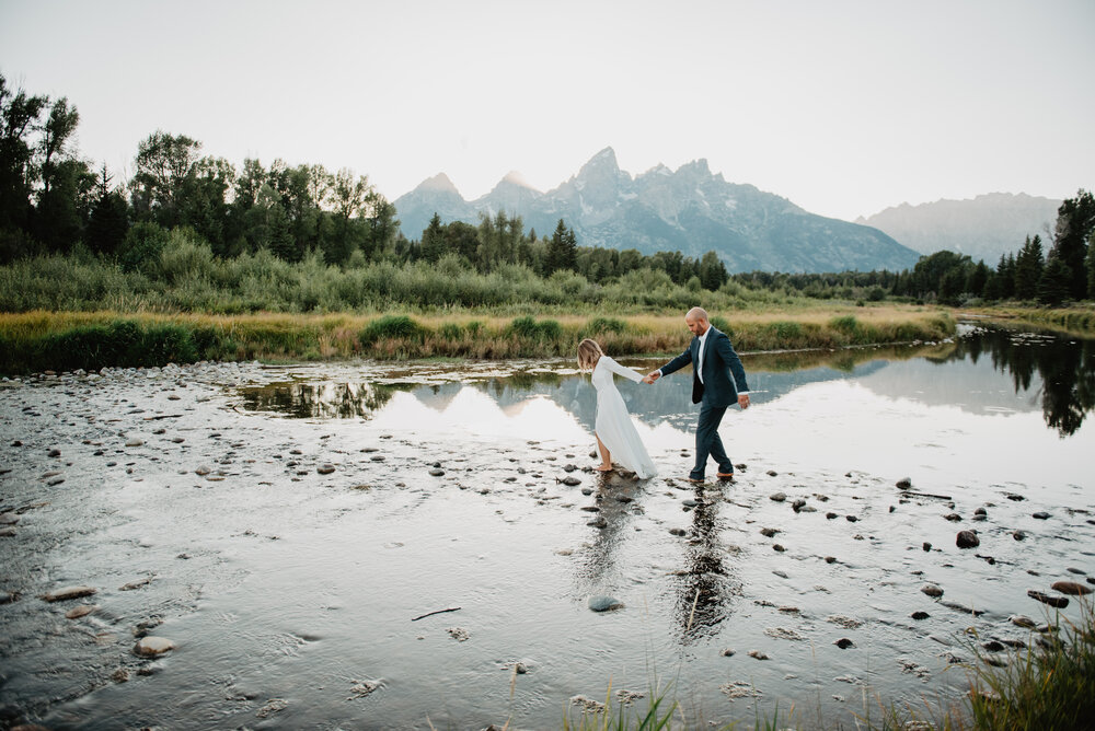 Grand Tetons elopement with bride holding the grooms hand and leading him through a stream in front of the Tetons Jocilyn Bennett Photography | How to Plan an Elopement | Why Elope | Reasons to Elope | Destination Wedding Photographer | Utah Wedding Photographer | Adventure Photographer | Destination Elopement Photographer | Traveling Photographer | Grand Teton Elopement | Grand Teton Photographer | National Park Weddings | Mountain elopement |