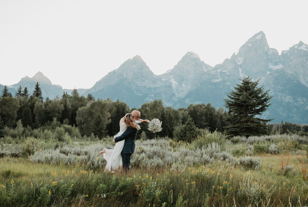 bride in a white flowy gown jumps on her groom in a sage brush field in Jackson Hole with the Tetons behind them and her groom holds her as they look out to the distance Jocilyn Bennett Photography | How to Plan an Elopement | Why Elope | Reasons to Elope | Destination Wedding Photographer | Utah Wedding Photographer | Adventure Photographer | Destination Elopement Photographer | Traveling Photographer | Grand Teton Elopement | Grand Teton Photographer | National Park Weddings | Mountain elopement |
