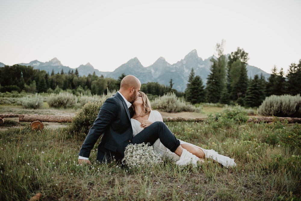 adventure elopement in the Grand Tetons with bride and groom sitting in a field in front of the mountains kissing with the brides babies breathe wedding bouquet at their feet Jocilyn Bennett Photography | How to Plan an Elopement | Why Elope | Reasons to Elope | Destination Wedding Photographer | Utah Wedding Photographer | Adventure Photographer | Destination Elopement Photographer | Traveling Photographer | Grand Teton Elopement | Grand Teton Photographer | National Park Weddings | Mountain elopement |