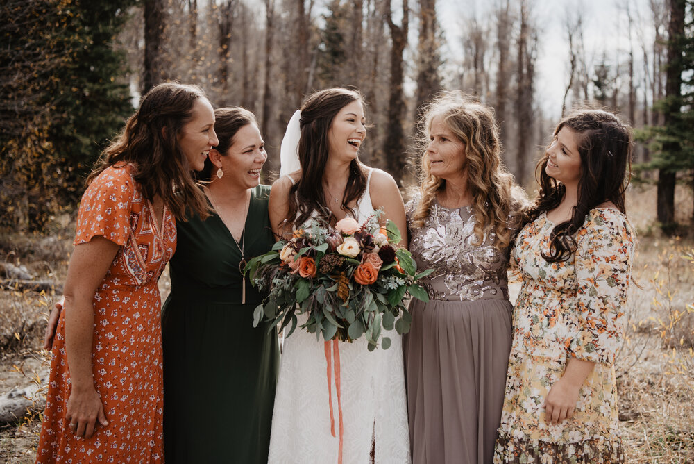 bride standing with her bridesmaids and mother in the woods in Jackson hole as they all laugh  together on her wedding day Jocilyn Bennett Photography | How to Plan an Elopement | Why Elope | Reasons to Elope | Destination Wedding Photographer | Utah Wedding Photographer | Adventure Photographer | Destination Elopement Photographer | Traveling Photographer | Grand Teton Elopement | Grand Teton Photographer | National Park Weddings | Mountain elopement |