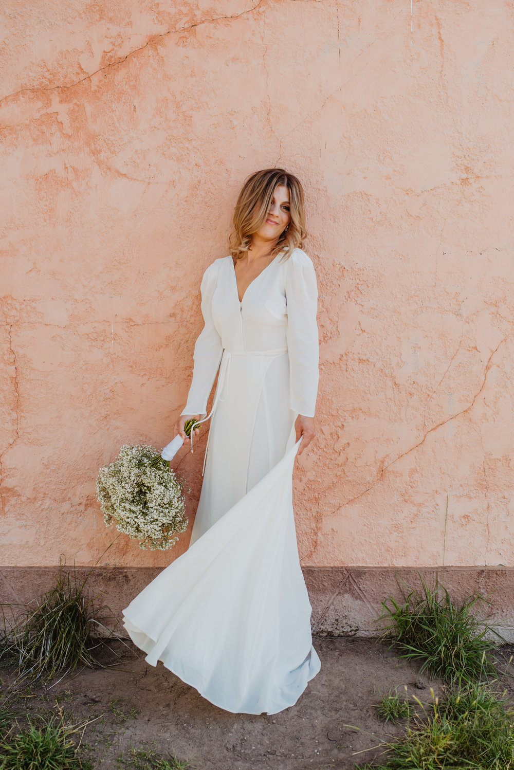 beautiful bride in a simple long sleeve white wedding dress and a babies breathe bouquet standing in front of a pinkish stucco wall for her adventure elopement Jocilyn Bennett Photography | How to Plan an Elopement | Why Elope | Reasons to Elope | Destination Wedding Photographer | Utah Wedding Photographer | Adventure Photographer | Destination Elopement Photographer | Traveling Photographer | Grand Teton Elopement | Grand Teton Photographer | National Park Weddings | Mountain elopement |
