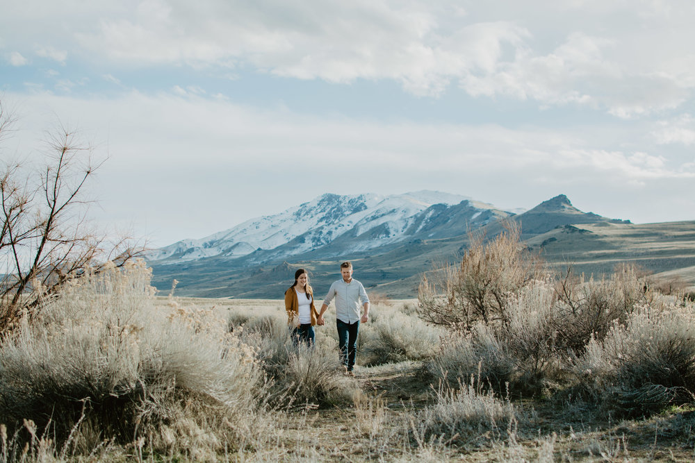 Antelope Island engagement session with man holding womans hand and leading her through a field full of desert brush with a mountain range behind them Jocilyn Bennett Photography | Destination Wedding Photographer | Elopement Wedding Photographer | National Park Elopement Photography | Capturing raw and genuine emotion | Utah Photographer | Utah wedding Photographer | National Park Weddings | Adventure Photographer | Antelope Island Engagements | Engagement pose ideas | Utah engagement photography | Engagement Pose Ideas | Candid Engagements |