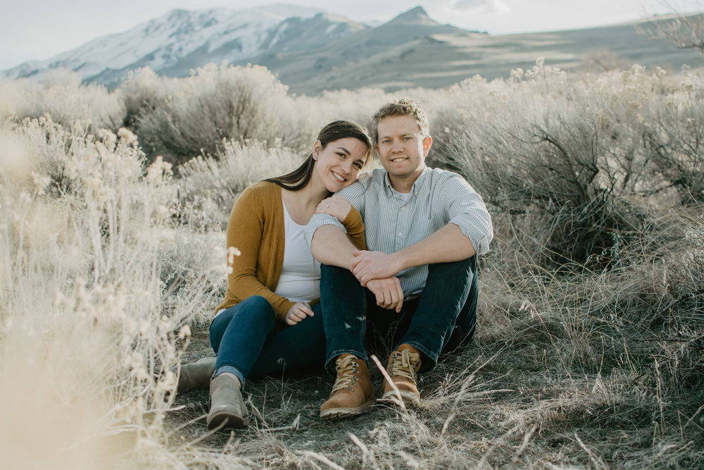 engaged couple sitting in a sage brush field in Antelope Island for their engagement session, with the woman wearing yellow and the man wearing grey, and mountains in the background Jocilyn Bennett Photography | Destination Wedding Photographer | Elopement Wedding Photographer | National Park Elopement Photography | Capturing raw and genuine emotion | Utah Photographer | Utah wedding Photographer | National Park Weddings | Adventure Photographer | Antelope Island Engagements | Engagement pose ideas | Utah engagement photography | Antelope Island | Candid Engagements | Antelope Island Photography |
