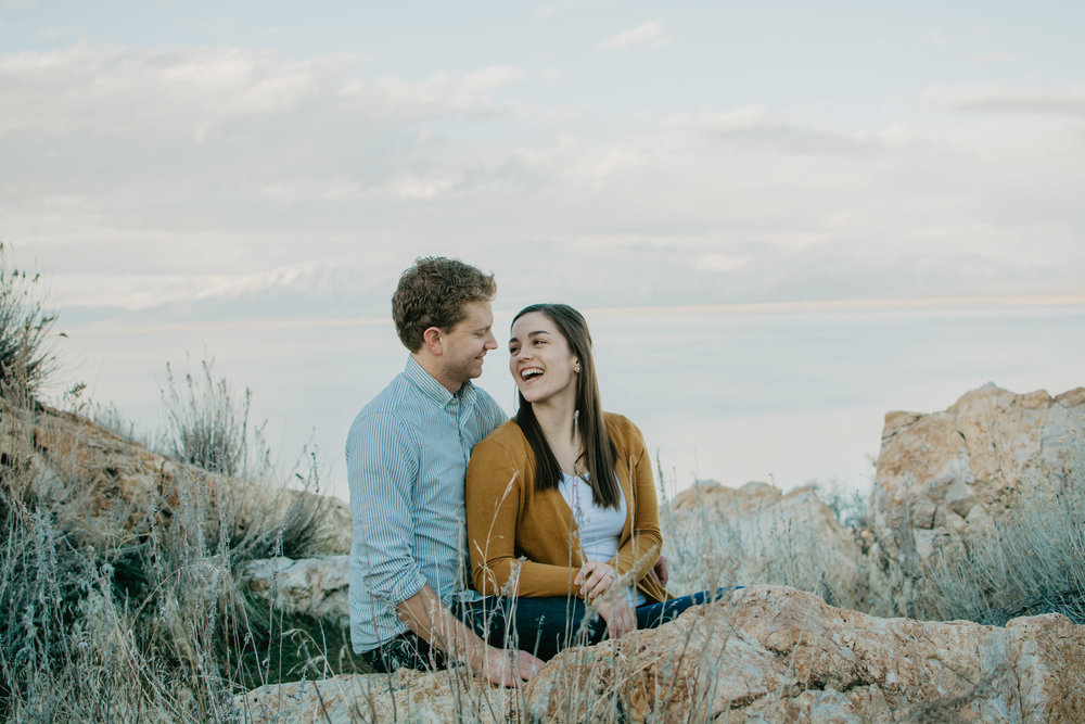 man and woman sitting between some boulder in Utah for their engagement session, the woman is looking at her fiance and laughing with a mountain in the back ground Jocilyn Bennett Photography | Destination Wedding Photographer | Elopement Wedding Photographer | National Park Elopement Photography | Capturing raw and genuine emotion | Utah Photographer | Utah wedding Photographer | National Park Weddings | Adventure Photographer | Antelope Island Engagements | Engagement pose ideas | Utah engagement photography | Engagement Pose Ideas | Candid Engagements |
