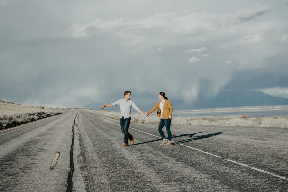Antelope Island engagement session with man holding a womans hand and walking across an empty road in Antelope Island as the thick clouds roll behind them Jocilyn Bennett Photography | Destination Wedding Photographer | Elopement Wedding Photographer | National Park Elopement Photography | Capturing raw and genuine emotion | Utah Photographer | Utah wedding Photographer | National Park Weddings | Adventure Photographer | Antelope Island Engagements | Engagement pose ideas | Utah engagement photography | Engagement Pose Ideas | Candid Engagements |
