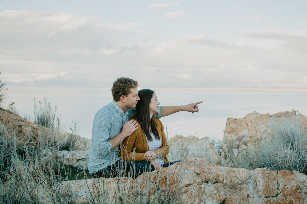 beautiful engagement photo of man holding woman by the shoulder and pointing into the distance as they sit on the top of a mountain and the couple smiles together Jocilyn Bennett Photography | Destination Wedding Photographer | Elopement Wedding Photographer | National Park Elopement Photography | Capturing raw and genuine emotion | Utah Photographer | Utah wedding Photographer | National Park Weddings | Adventure Photographer | Antelope Island Engagements | Engagement pose ideas | Utah engagement photography | Engagement Pose Ideas | Candid Engagements |