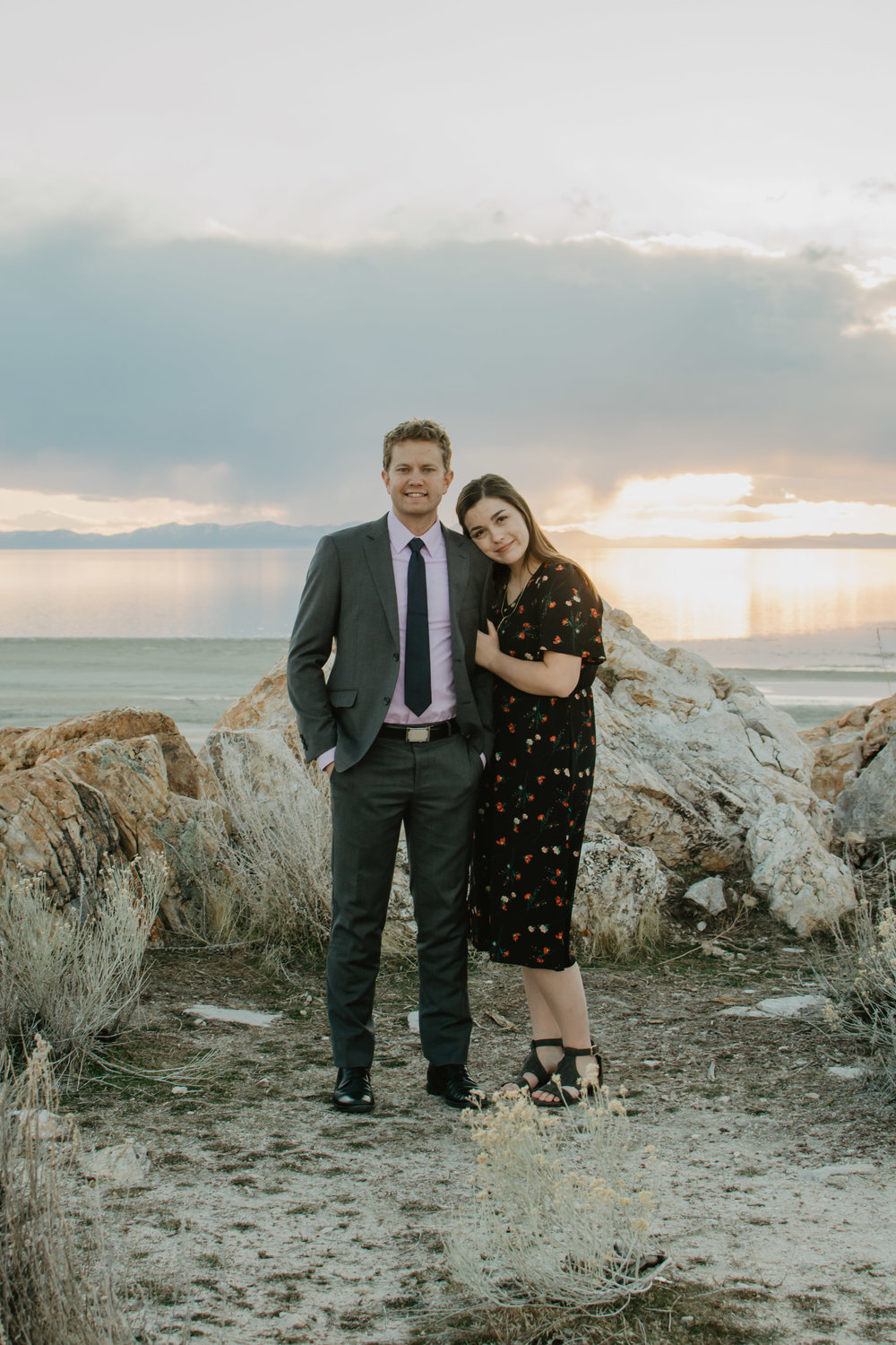 beautiful sunset engagement photo with thick rain clouds in the back ground with a sun set as the woman holds her fiances arm and they both smile at the camera Jocilyn Bennett Photography | Destination Wedding Photographer | Elopement Wedding Photographer | National Park Elopement Photography | Capturing raw and genuine emotion | Utah Photographer | Utah wedding Photographer | National Park Weddings | Adventure Photographer | Antelope Island Engagements | Engagement pose ideas | Utah engagement photography | Engagement Pose Ideas | Candid Engagements |