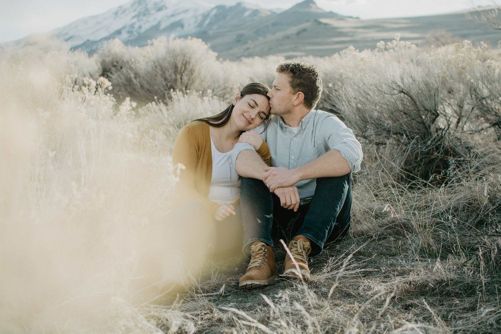 woman sitting with her fiance in a sage brush field in Antelope Island as she leans on his shoulder and holds his arm and he kisses her foreheadJocilyn Bennett Photography | Destination Wedding Photographer | Elopement Wedding Photographer | National Park Elopement Photography | Capturing raw and genuine emotion | Utah Photographer | Utah wedding Photographer | National Park Weddings | Adventure Photographer | Antelope Island Engagements | Engagement pose ideas | Utah engagement photography | Engagement Pose Ideas | Candid Engagements |