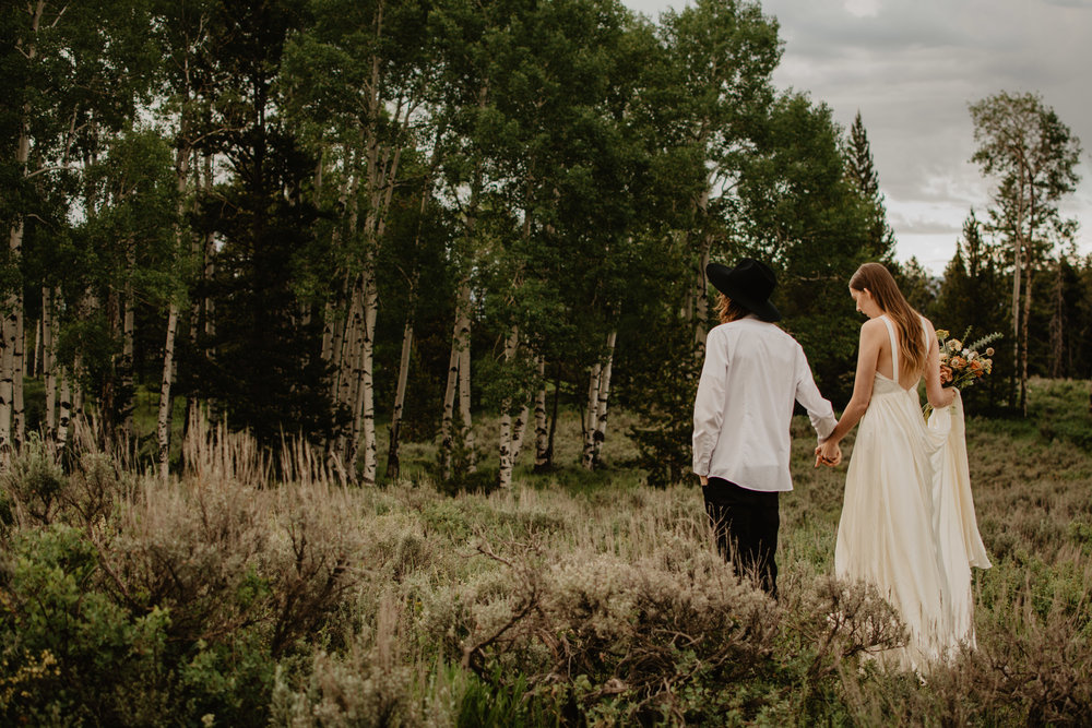 bride and groom holding hands and walking towards a wooded area for their jackson hole elopement photos