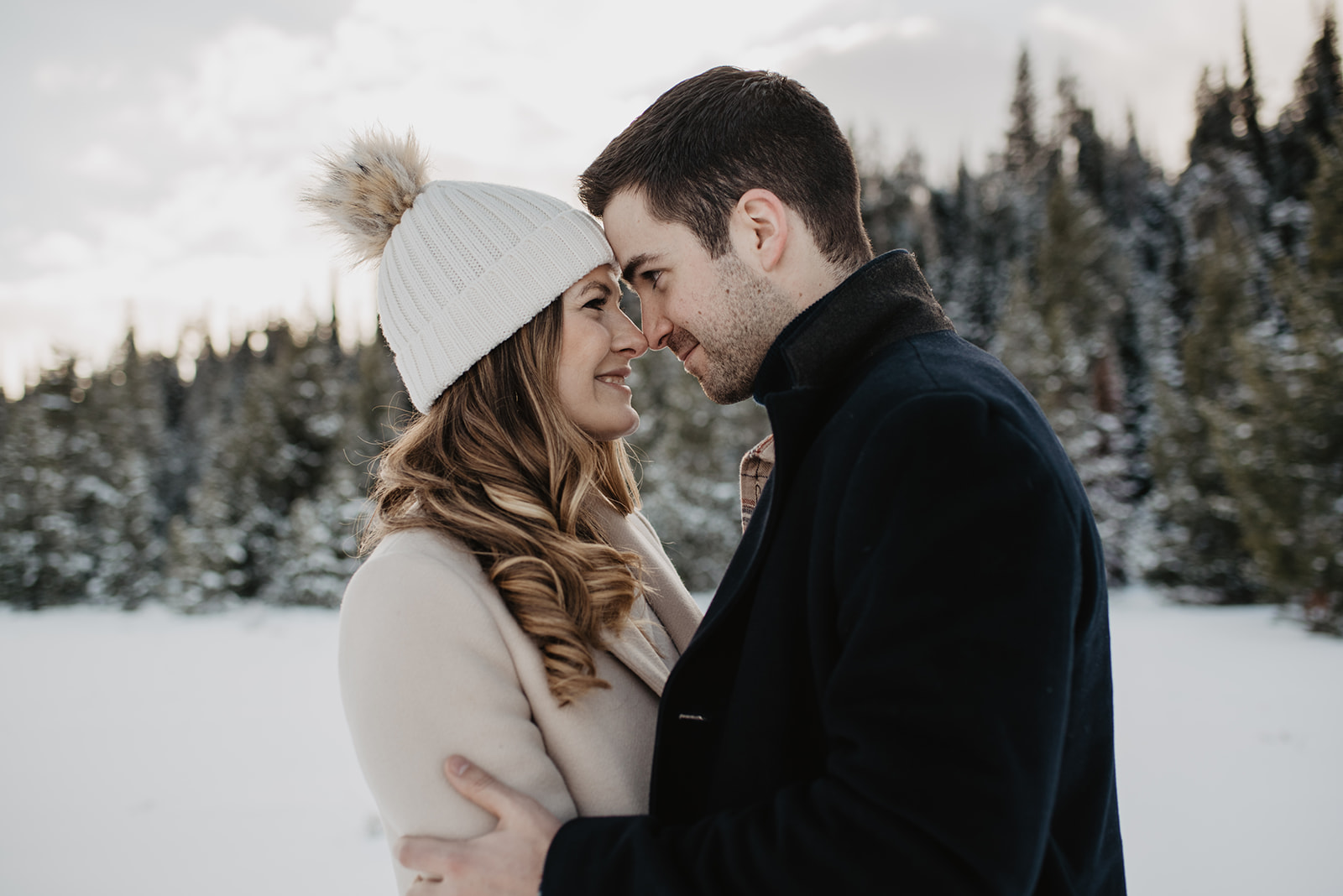 man holding woman by the shoulders with his forehead to her forehead as they smile at each other while standing in the winter snow in front of pine trees in Jackson Hole