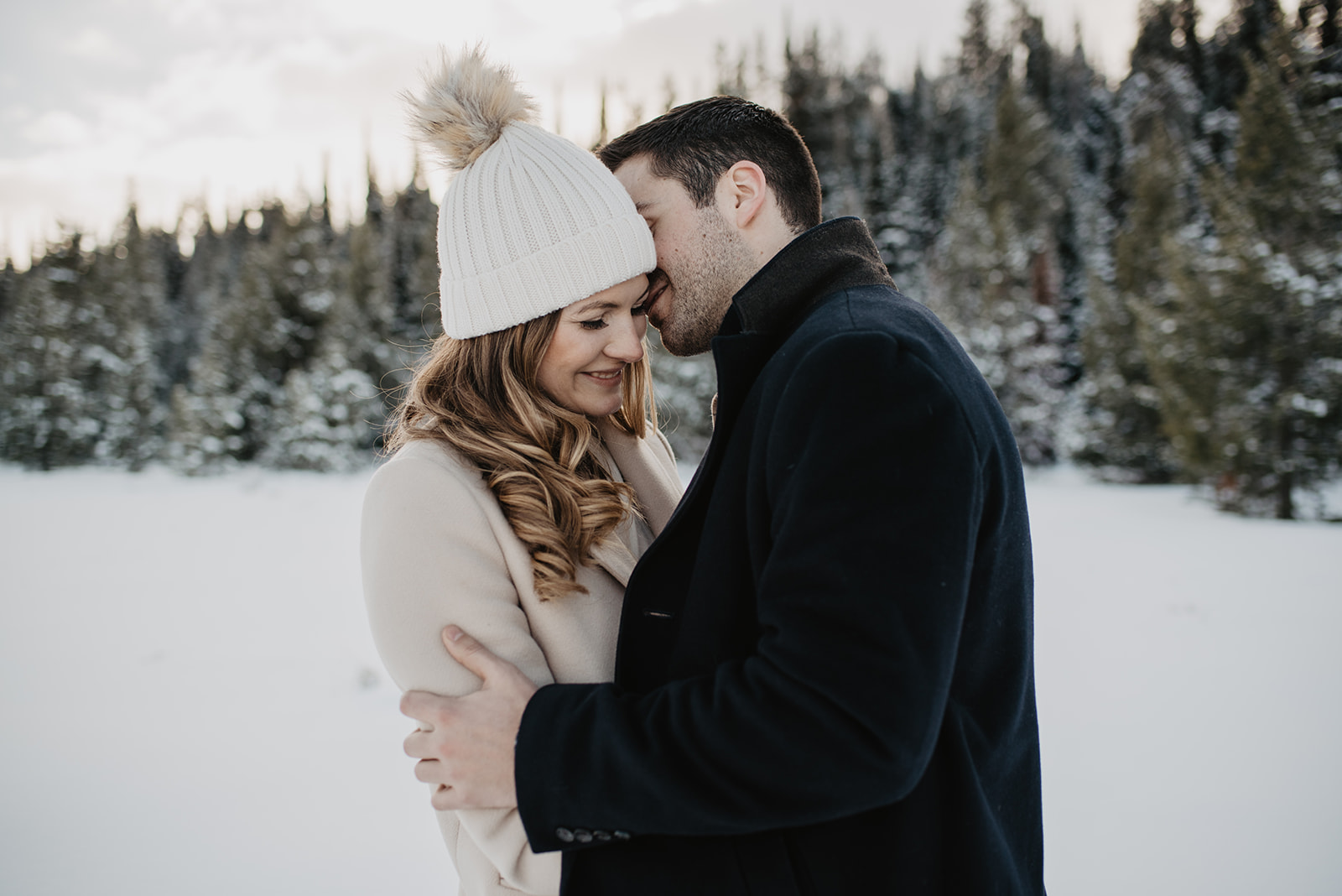 man whispering in woman's ear while holding her close as she looks down and smiles in the snow in front of pine tress in Jackson Hole