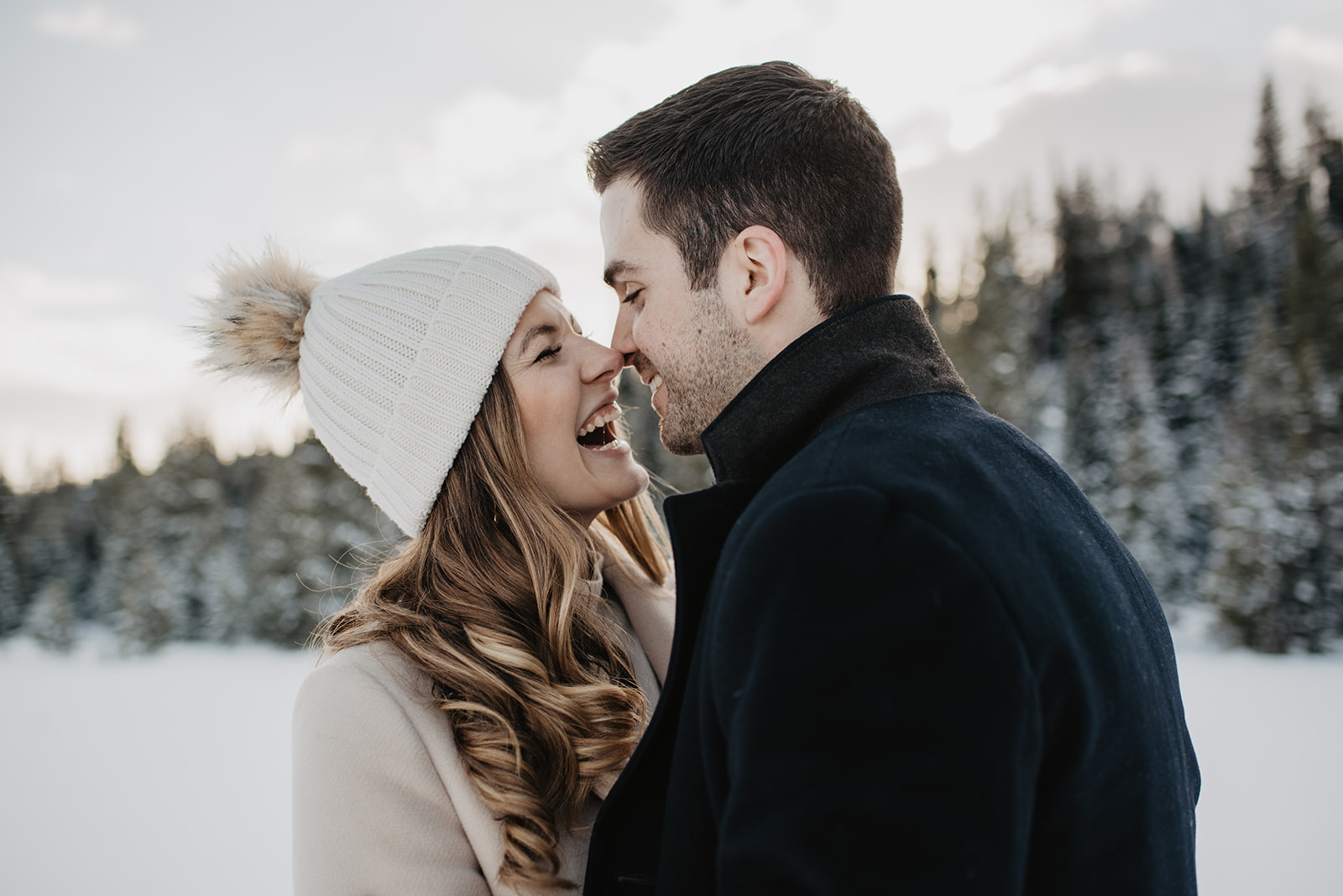 winter engagement session in Jackson Hole with man and woman laughing while they hold each other in fron tof pine tress and snow covered ground