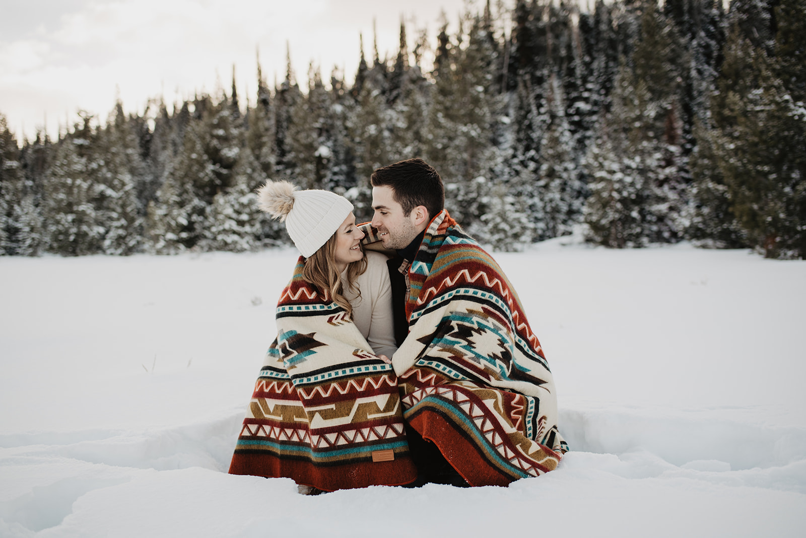 Winter engagement session with man and woman cuddled up in a wool tribal print blanket while sitting in a snowy field while cuddling and looking at each other with pine trees behind them in Jackson Hole