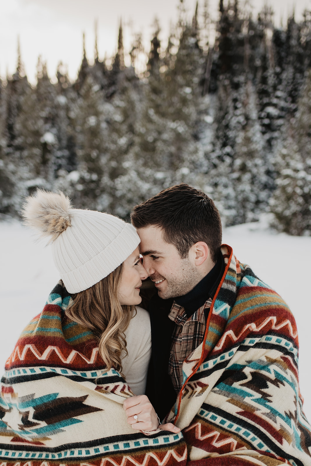 Jackson Hole winter engagement session with man and woman snuggling together in a tribal print wool blanket in the snow with big pine trees behind them