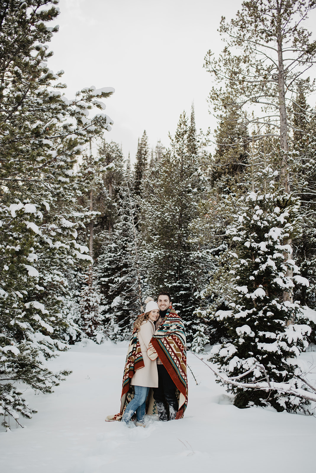 Jackson Hole winter engagement session with couple standing in a grove of pine trees wrapped in a wool tribal print blanket