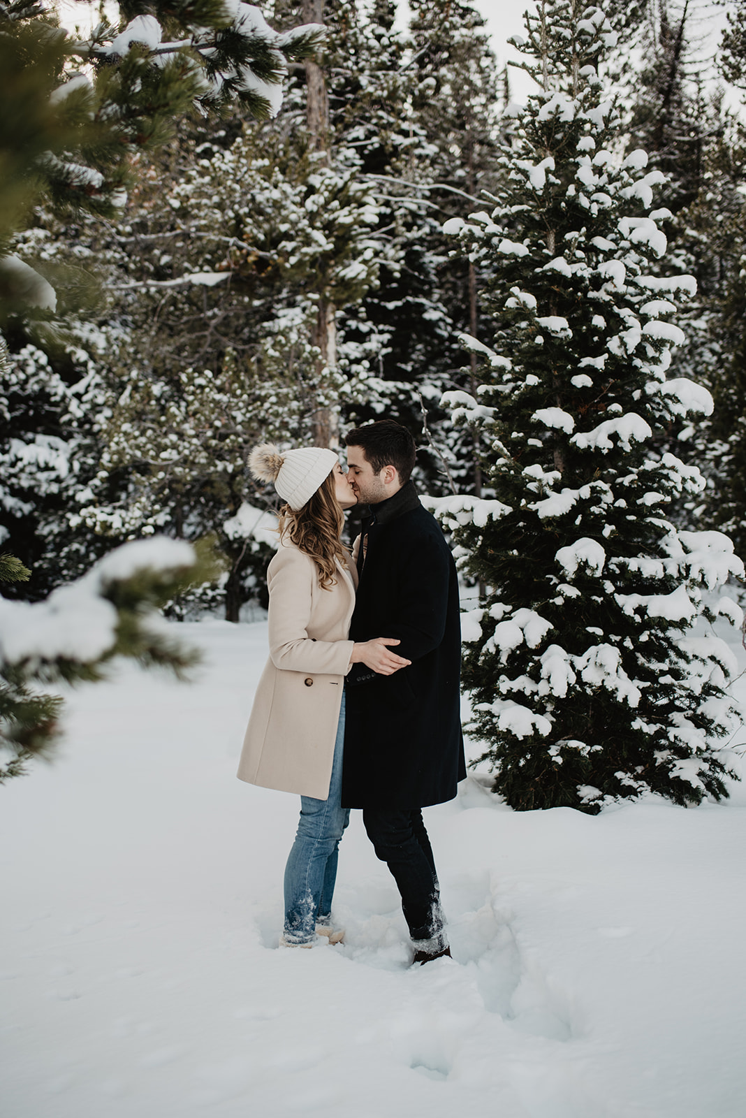 winter engagement session with man and woman standing while surrounded by pine trees as they cuddle up to each other