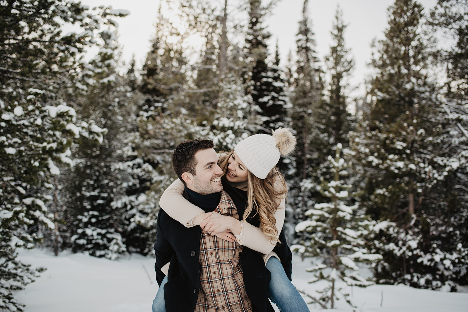 man holding a woman on his back as she looks over his shoulder smiling and looks at her as they stand in front of pine trees in Jackson Hole for their winter engagement session
