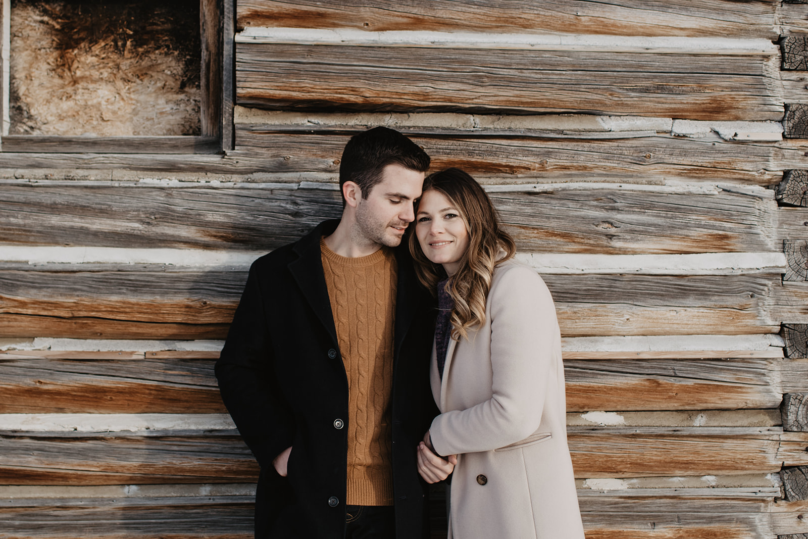 man and woman standing on a wooden barn wall while the man looks down at the woman and the woman looking into the camer and smiles in Jackson Hole for their winter engagement session