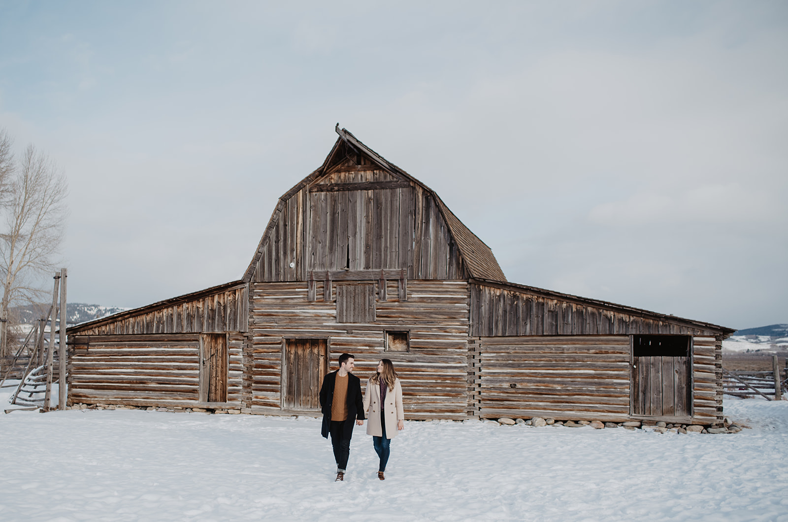 Man and woman walking towards the camera while holding hands in a snowy field with an old wooden barn behind them for their Jackson Hole winter engagement session
