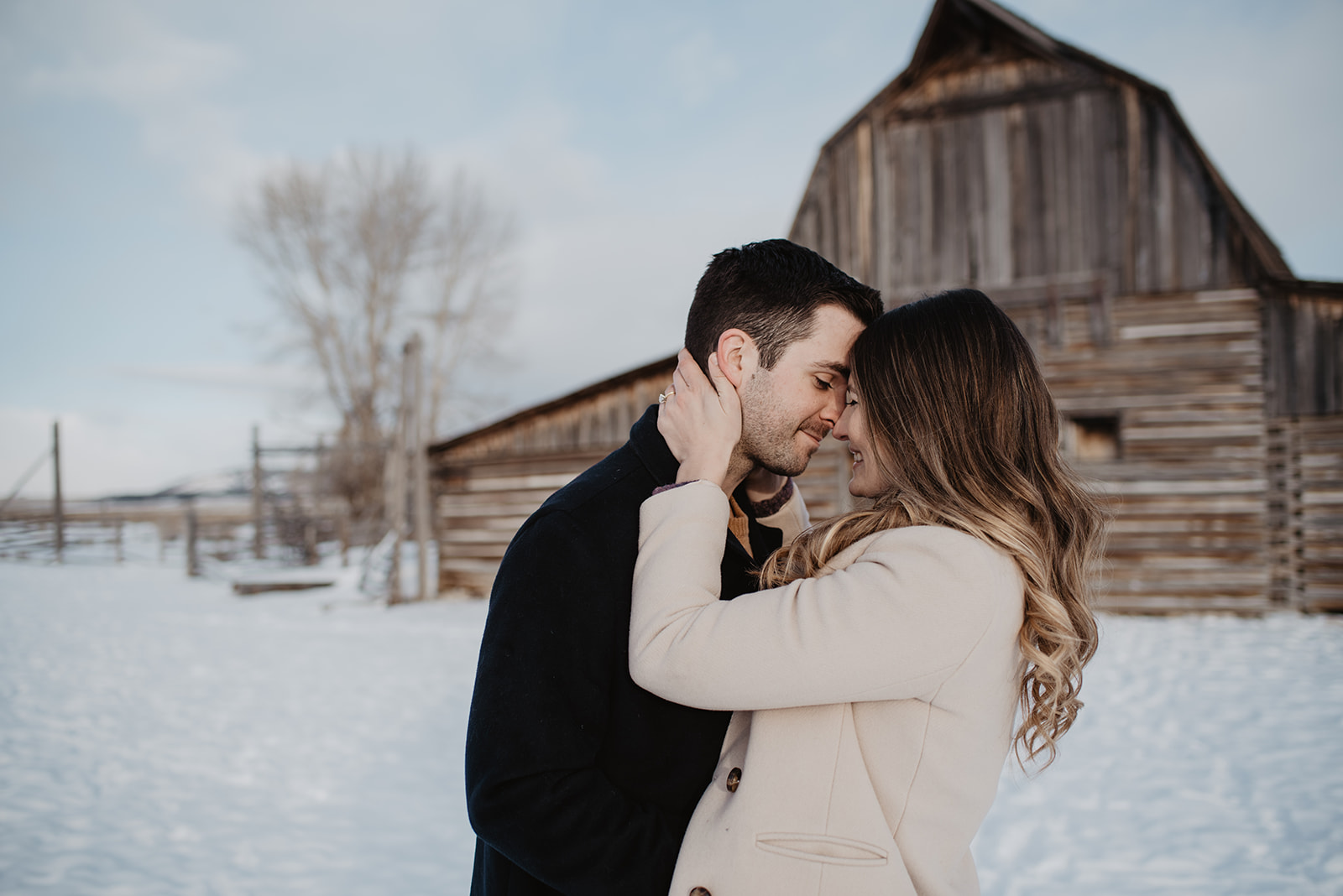 woman holding the back of man's neck and bringing he forehead to hers as they both smile in front of a wooden barn in Jackson Hole