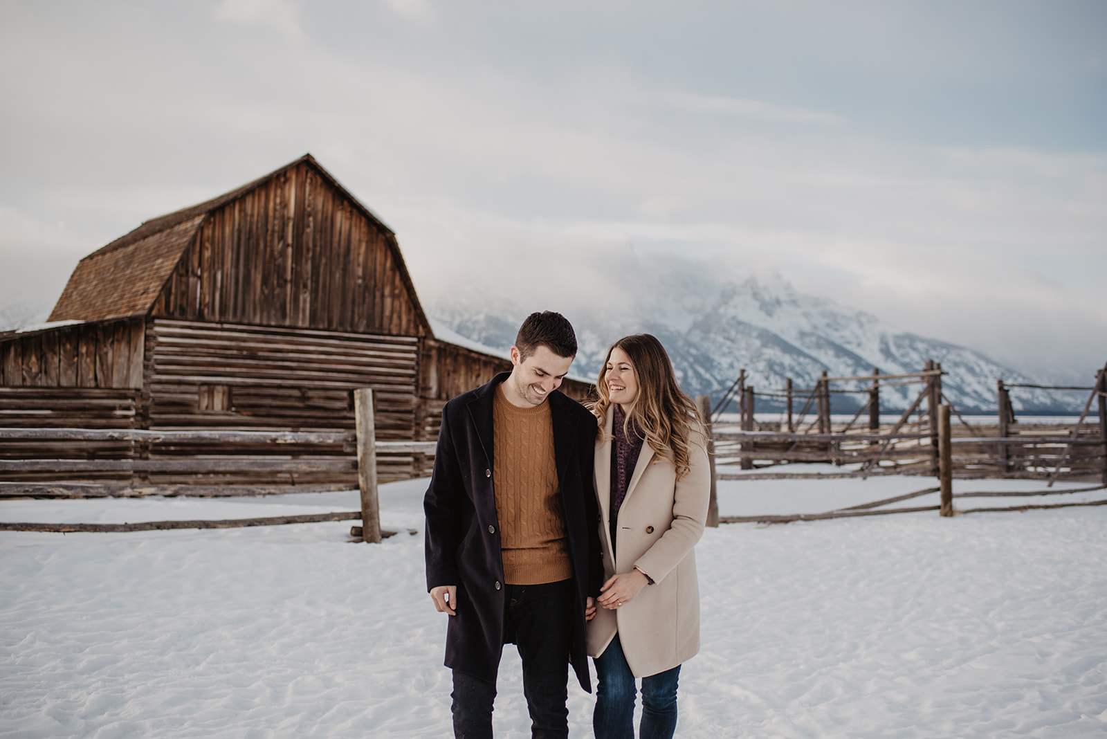 Jackson Hole winter engagement sessionwith couple walking in front of an old brown wooden bard in front of the Tetons while holding hands and dressed up