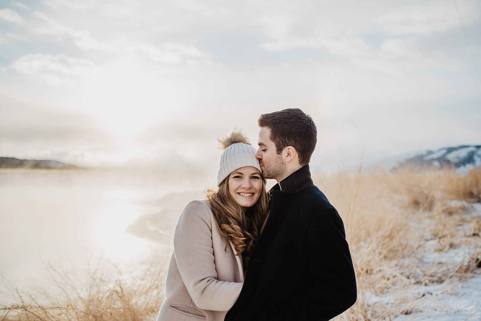Jackson Hole Winter Engagement Session in front of a lake in Jackson Hole as teh sun set and gleams behind the couple, the man is kissing the woman's forehead as she looks at the camera and smiles