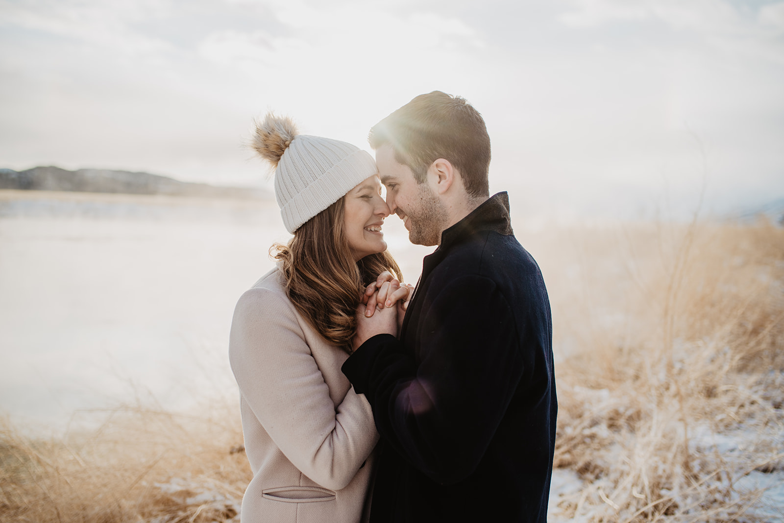 man and woman holding each others hands to their chests with their foreheads touching and smiling as the sun set behind them over a lake at Jackson Hole for their engagement session