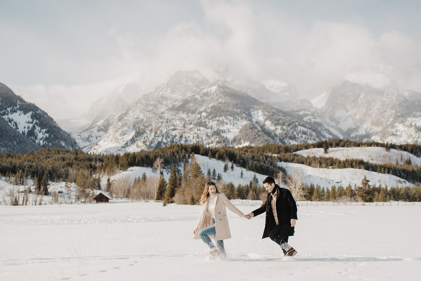 engaged couple walking through the snow in front of mountains in Jackson Hole for their winter engagement session while holding hands with the woman leading the man