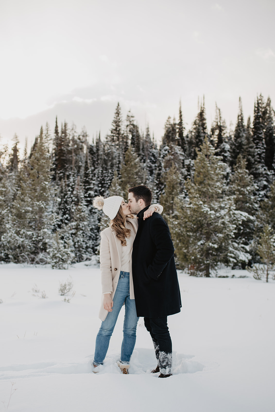 winter engagement session in Jackson HOle with couple standing in the snow with the womand arms aroudn teh mans neck and kissing him as they stand in front of tall pines