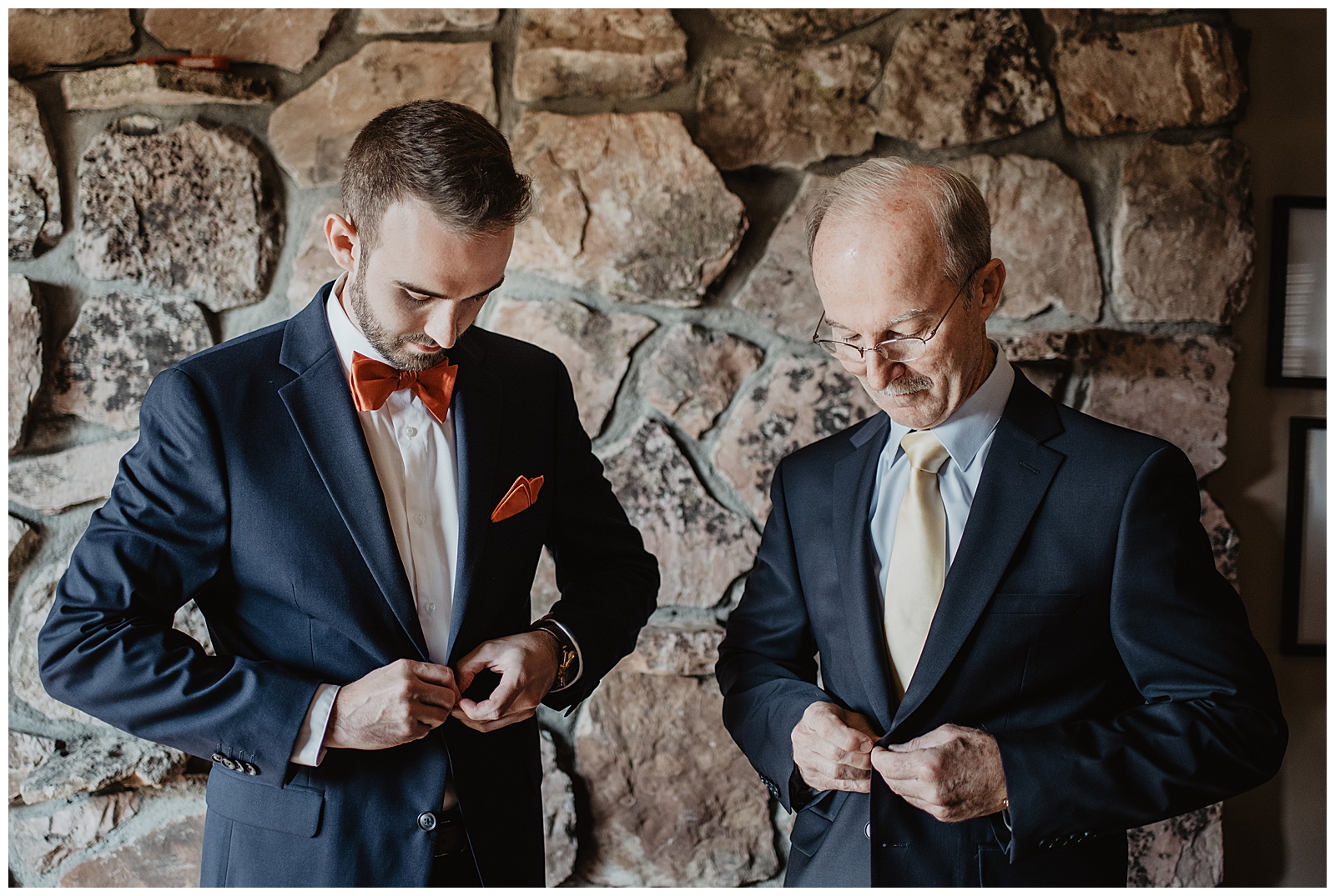 groom and his father buttoning their suit coats together while standing in front of a stone wall in the Tetons for the grooms fall wedding day