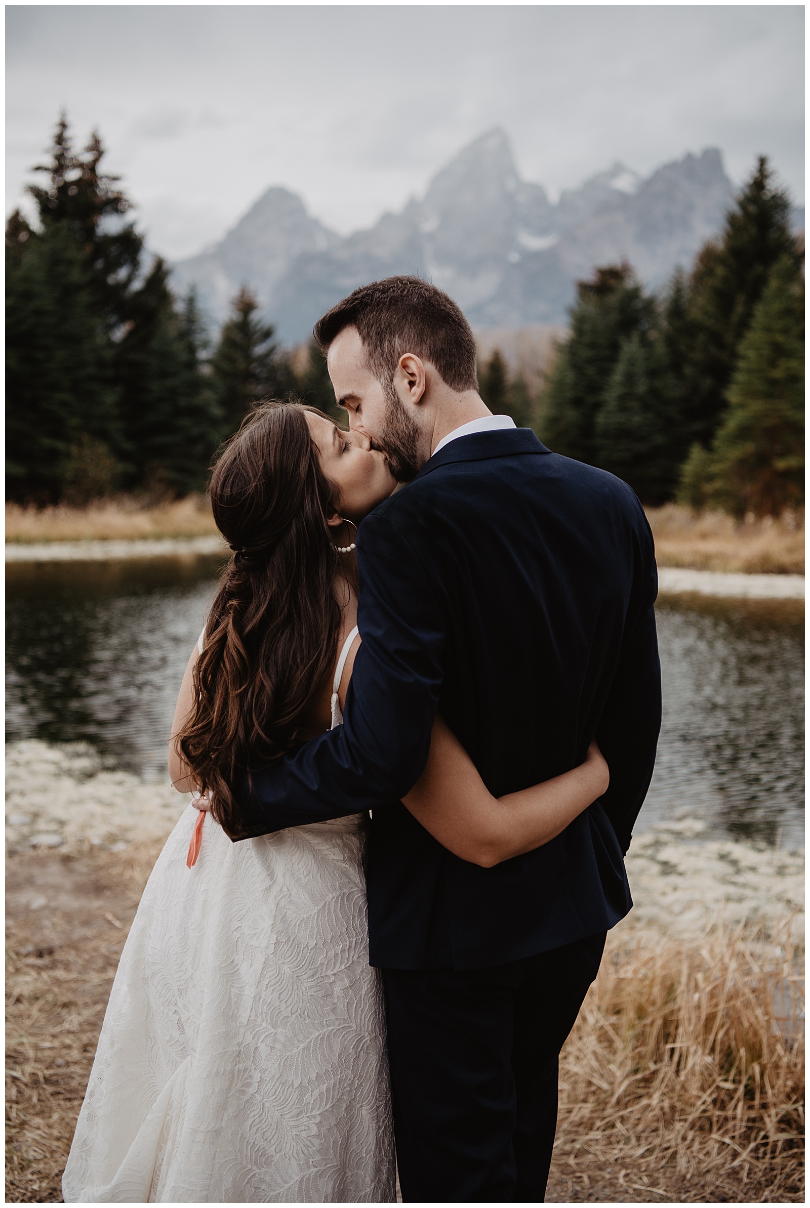 Grand Teton National Park fall weddingwith bride and groom holding each other by the waist and kissing in front of the Tetons with a creek near them