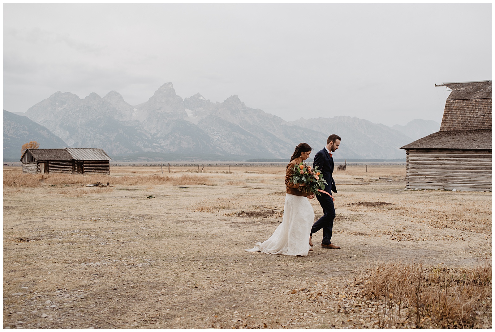bride wearing her wedding dress and a brown coat while holding her wedding bouquet, she is holding her grooms hand while walking across a dead field in front of The Grand Tetons on their fall wedding day