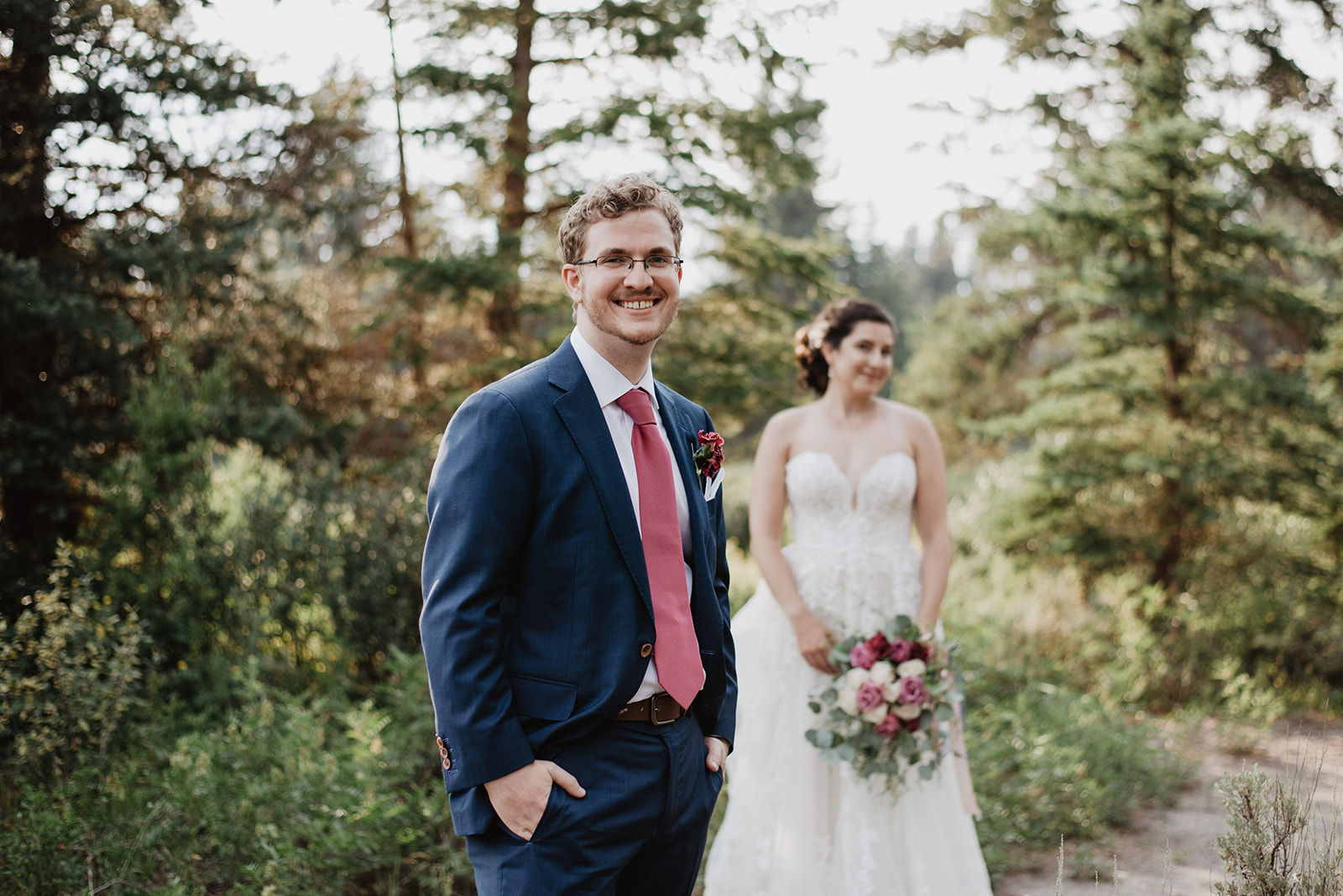 wedding photo of groom in a blue suit and red tie in Jackson Hole Wyoming smiling at the camera with his bride behind him and out of focus