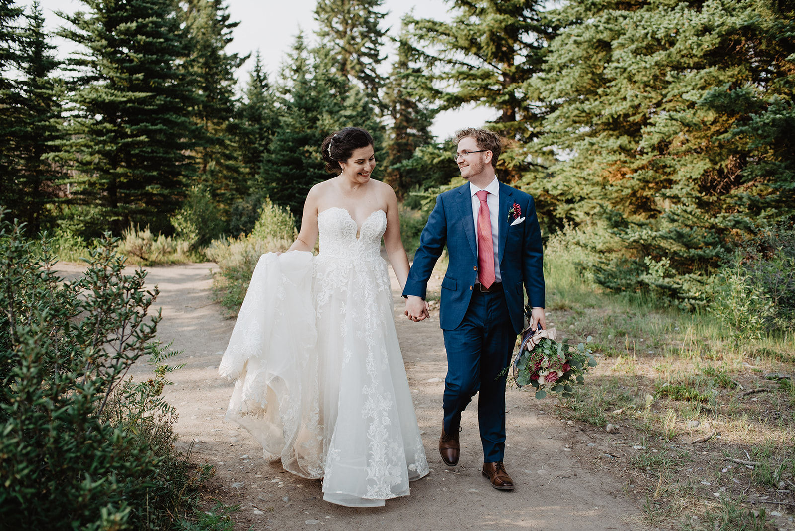 beautiful bride and groom walking on a dirt trail in Jackson Hole at Trail Creek Ranch as the bride holds her lace wedding dress up off the ground and her groom stares at her beauty