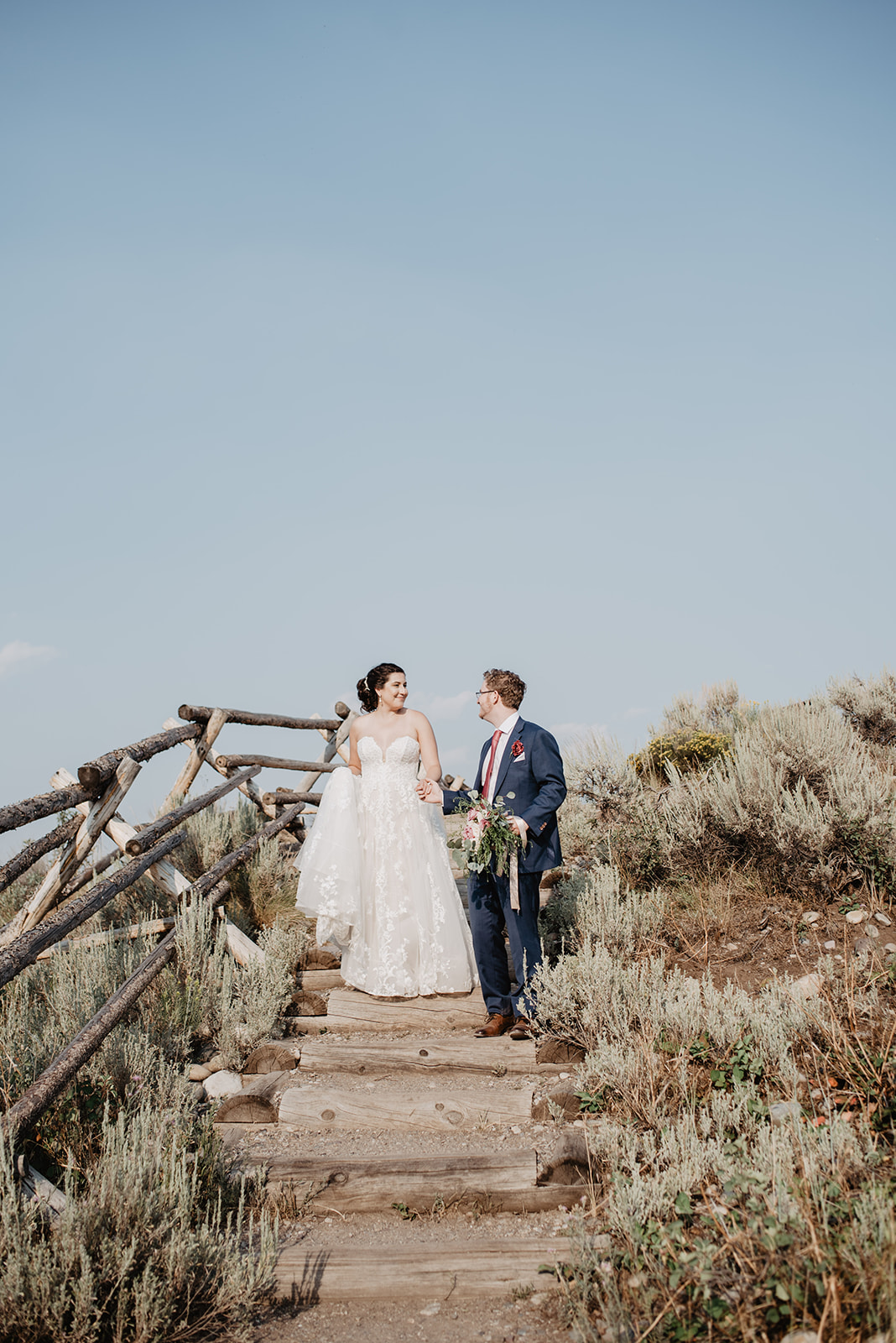 bride and groom walking down a wood and dirt stairway in the Tetons with sage brush around them and blue skies behind them. Bride is in a dainty lace dress with a full skirt
