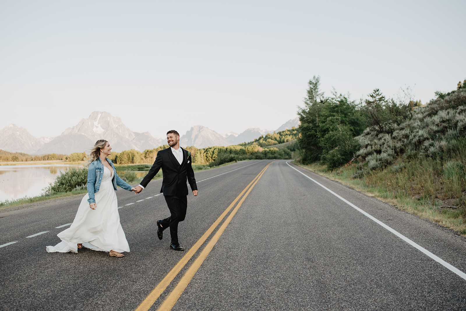 Jackson Hole wedding day at Diamong Cross ranch with groom holding his brides hand as they cross a road in front of the Grand Tetons. Bride is wears a white full gown with a denim jacket over the top and the groom is wearing a classic black suit