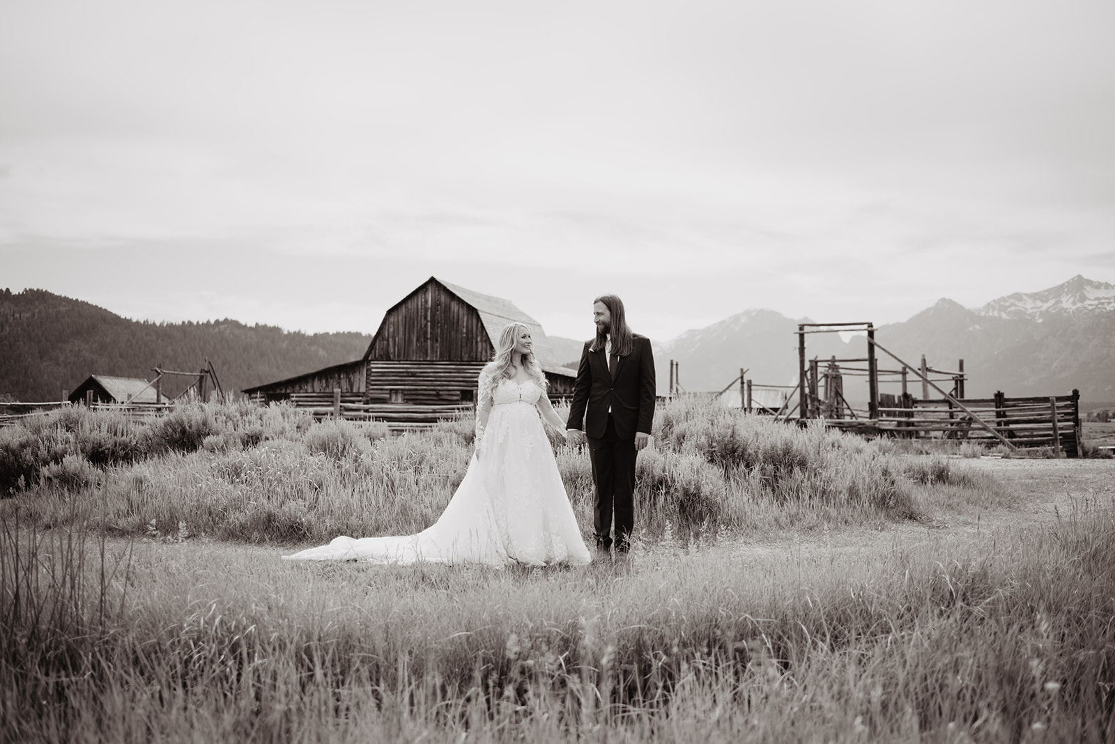 stunning black and white photo of bride in a flowy and full white gown holding her groom hands as they standing in front of a large barn in the Grand Tetons with mountains behind them